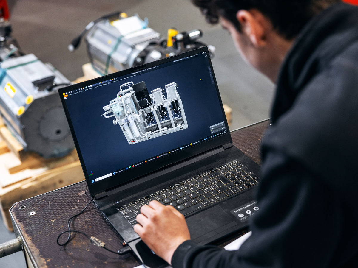 HydTec engineer reviewing a 3D CAD model of the hydraulic power unit assembly on a laptop, with physical pump components visible in the background