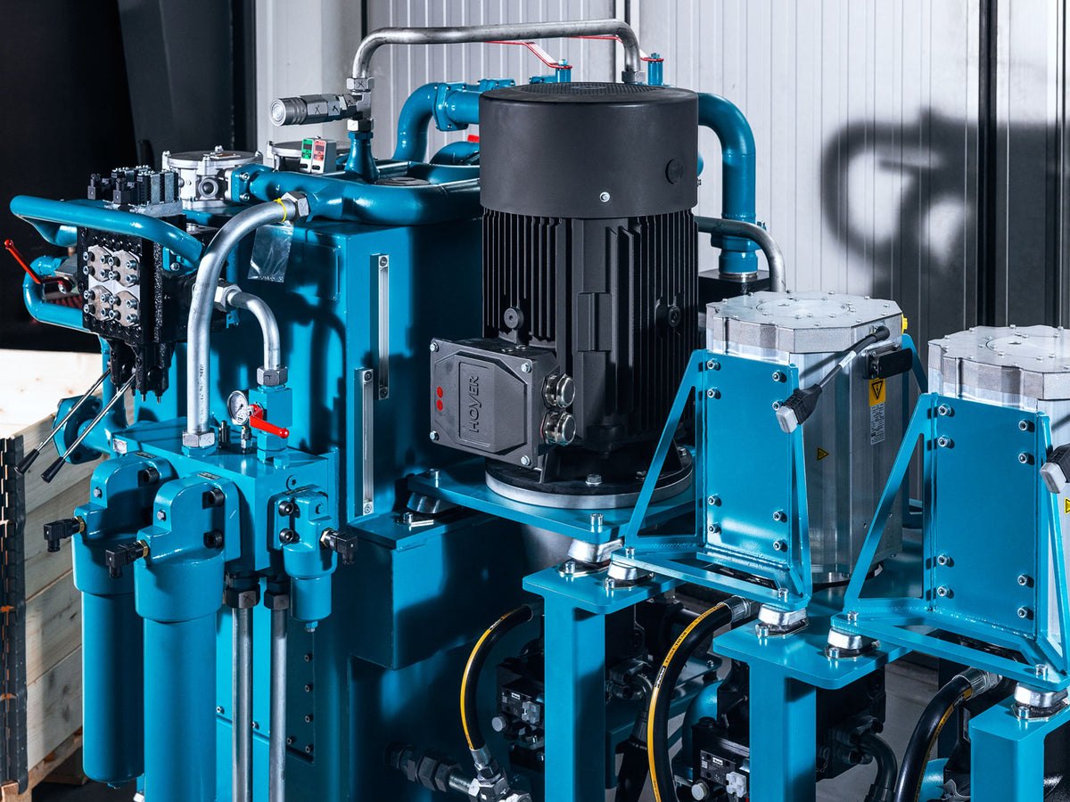 Industrial hydraulic power unit with a prominent black electric motor, blue metal housing, silver filter cylinders, and piping, photographed in a workshop setting.