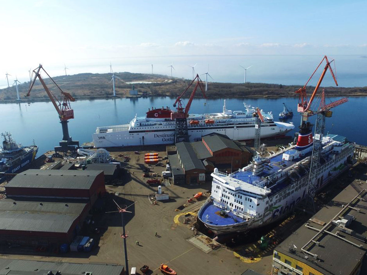 Aerial view of a shipyard harbor with multiple large passenger ferries in dry dock, red cranes, industrial buildings, and a distant shoreline with wind turbines under a pale blue sky.