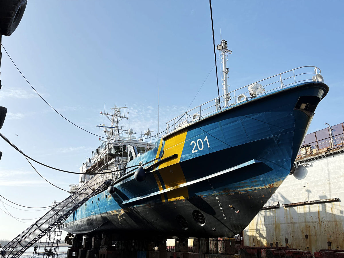 A large blue and yellow industrial ship or vessel photographed in a dry dock, with the hull number 201 visible on the front. The ship is supported by supports on the ground, with another ship and dock structures in the background under a clear blue sky.