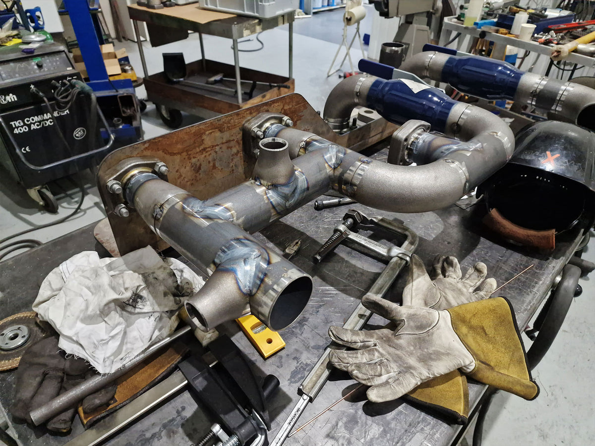 A set of custom welded metal exhaust pipes with blue and silver sections, displayed on a workbench in a workshop alongside welding gloves, a helmet, and tools.