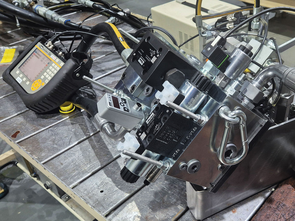 Industrial hydraulic control valve assembly with metal cylinders, hoses, wiring, and a handheld diagnostic tool on a workbench.