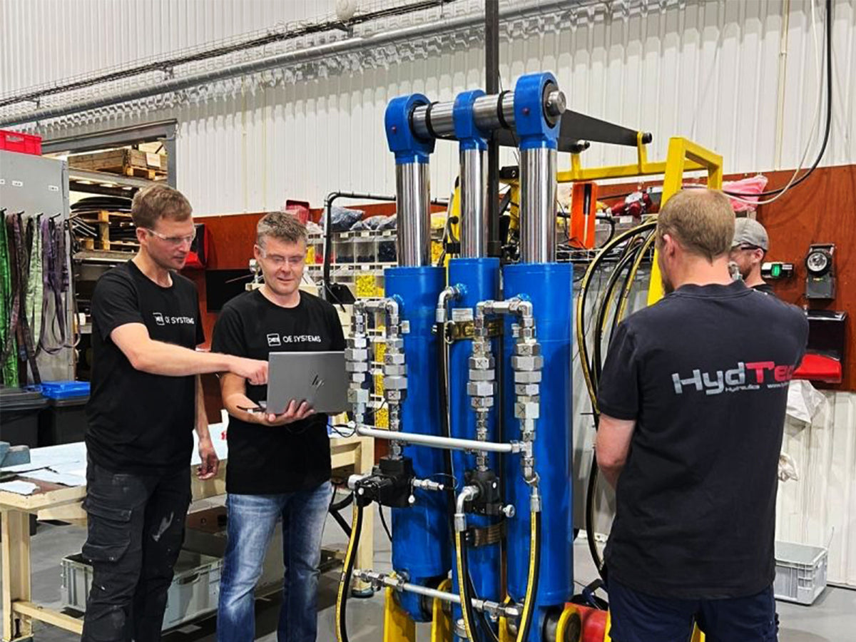 Two hydtec employees and two OE Systems employees stand beside a large blue and yellow industrial hydraulic machine in a workshop, with one holding a laptop.