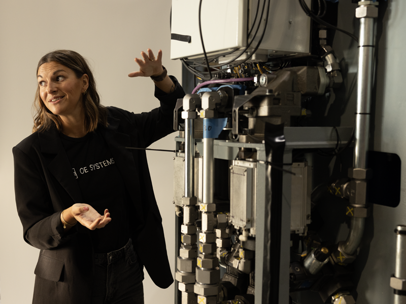 A woman in a black blazer and t-shirt stands next to a large industrial boiler or heat pump system, gesturing toward the machinery. The t-shirt features the text 'OE SYSTEMS'.