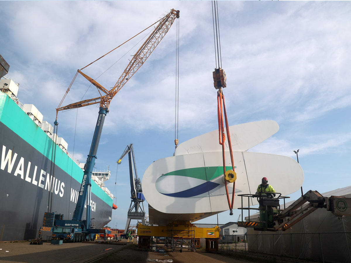 A large white metal section with a green and blue wave logo is being lifted by a crane in a shipyard, next to a teal and black vessel labeled WALLENIUS.