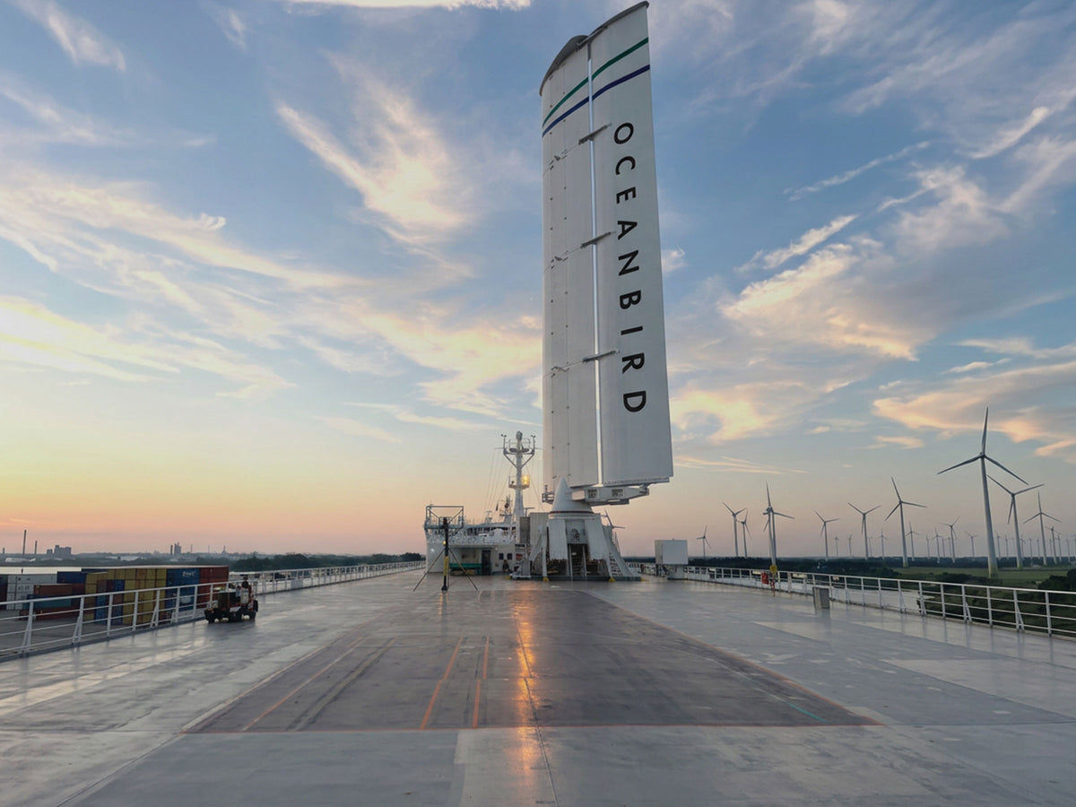 Oceanbird's full-scale wing sail on a vessel deck at Oresund DryDocks in Landskrona, wind turbines visible at sunset.