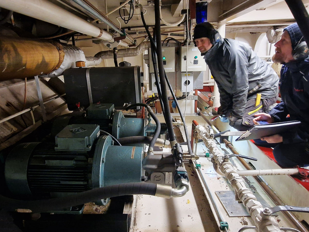 Two men inspecting industrial machinery and piping inside a confined technical space, with one holding a tablet.