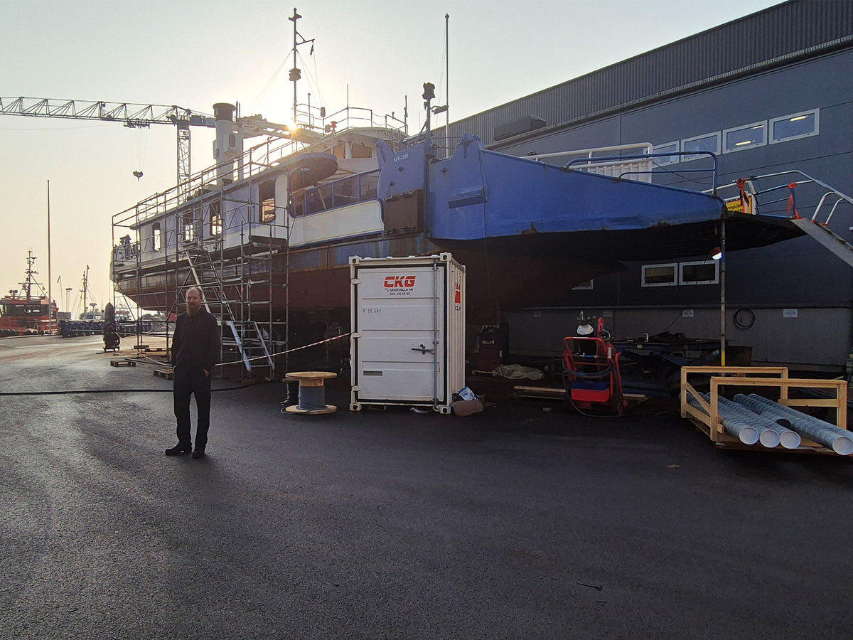 A large blue and dark gray boat or ferry under construction is parked outdoors beside a building and crane, with scaffolding around the hull. A person stands on the wet pavement in the foreground, and construction equipment are visible.