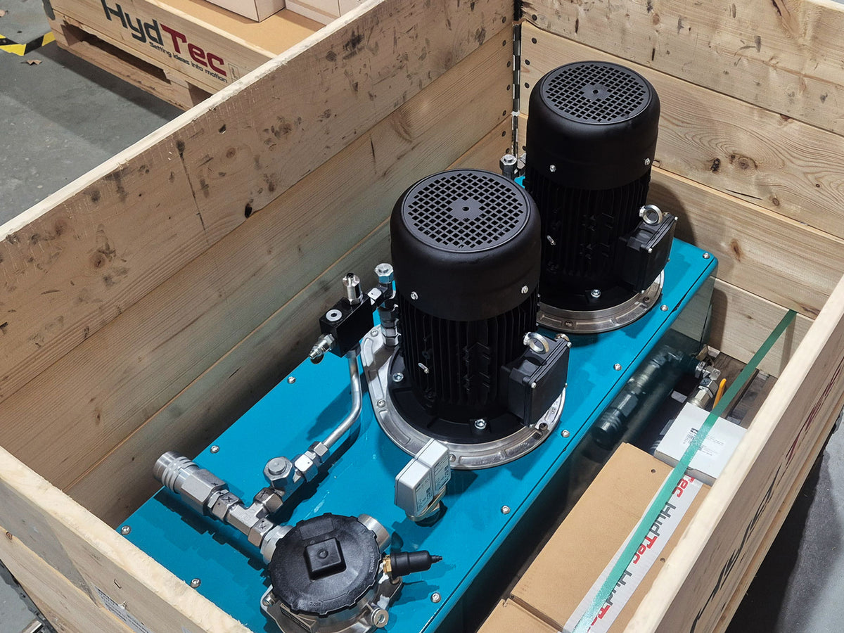 A HydTec industrial fluid power pump system with two black motors and a blue metal base, packaged inside a wooden shipping crate.