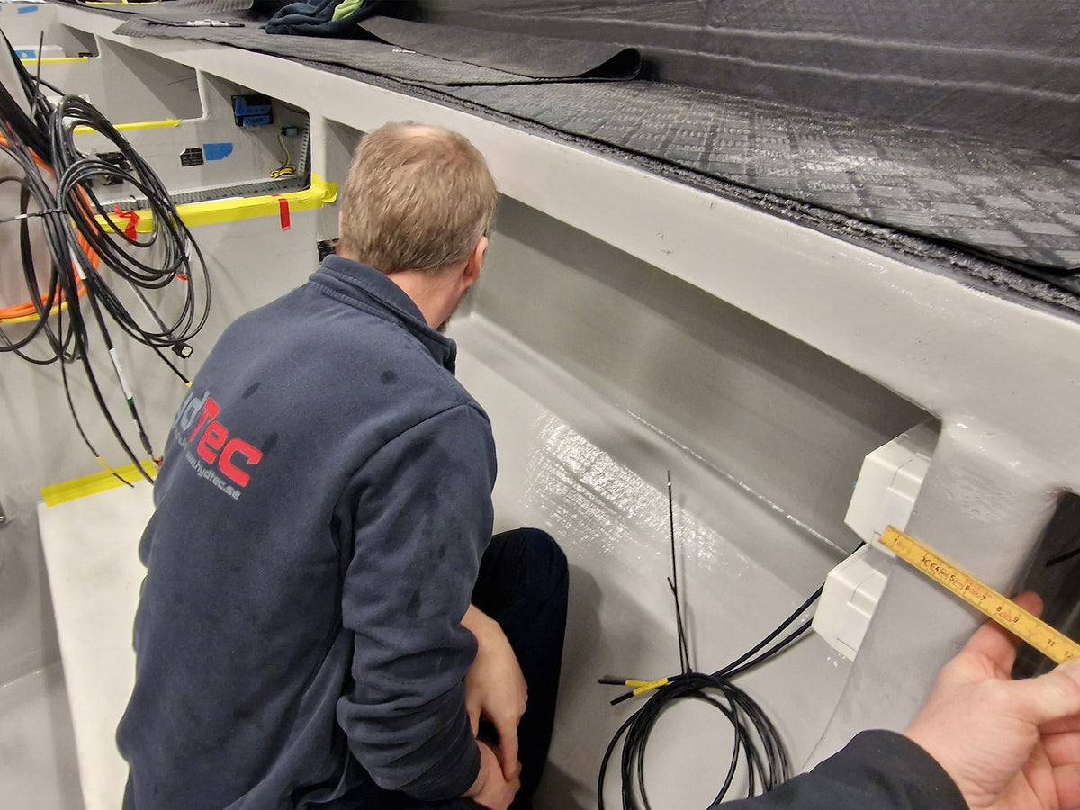 A man in a navy blue work jacket with a Hydtec logo on the back kneels inside a white fiberglass boat cabin. A second person is measuring a fitting with a yellow tape measure. Black wiring is visible on the floor of the cabin.