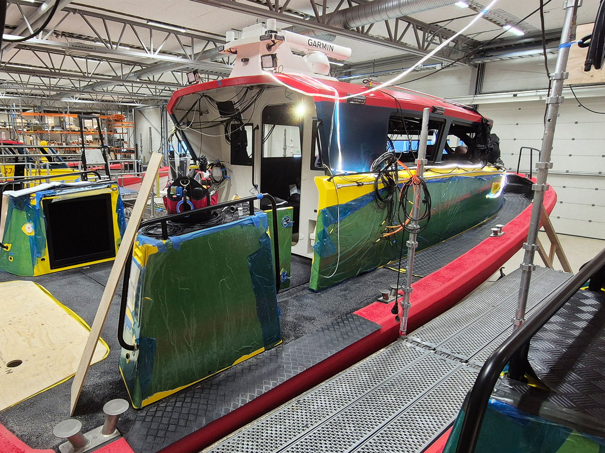 A red, yellow, and green coast guard boat under construction inside a workshop, with a metal walkway leading alongside it.