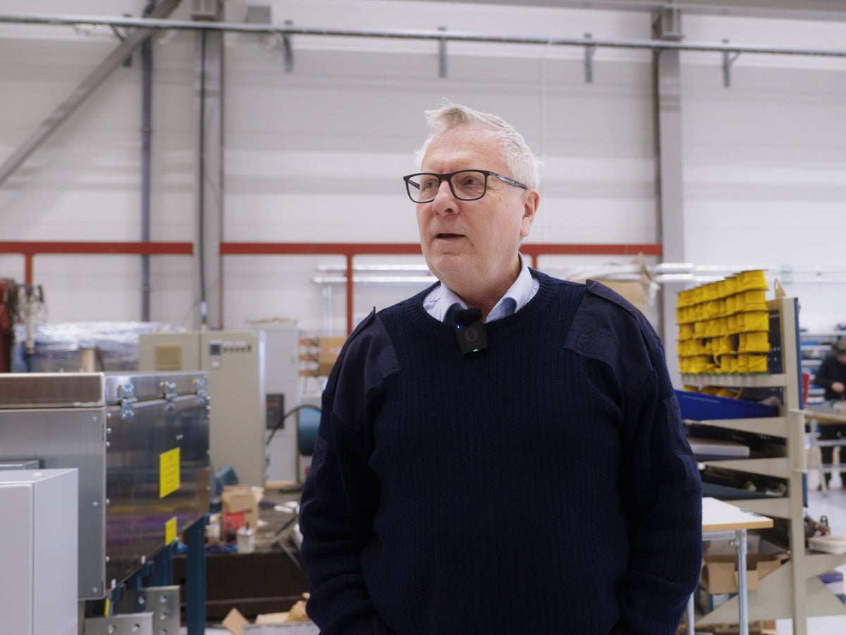 A man with white hair and glasses, wearing a dark navy sweater over a light blue collared shirt, standing in a large industrial workshop.