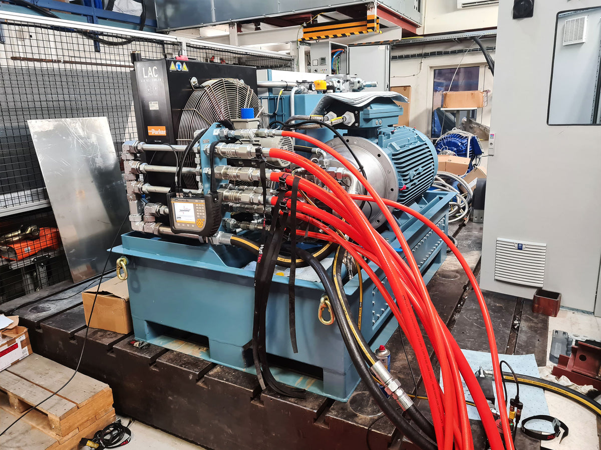 A large industrial hydraulic power unit with a blue base, electric motor, and multiple red and black hydraulic hoses, photographed in a workshop or factory setting.