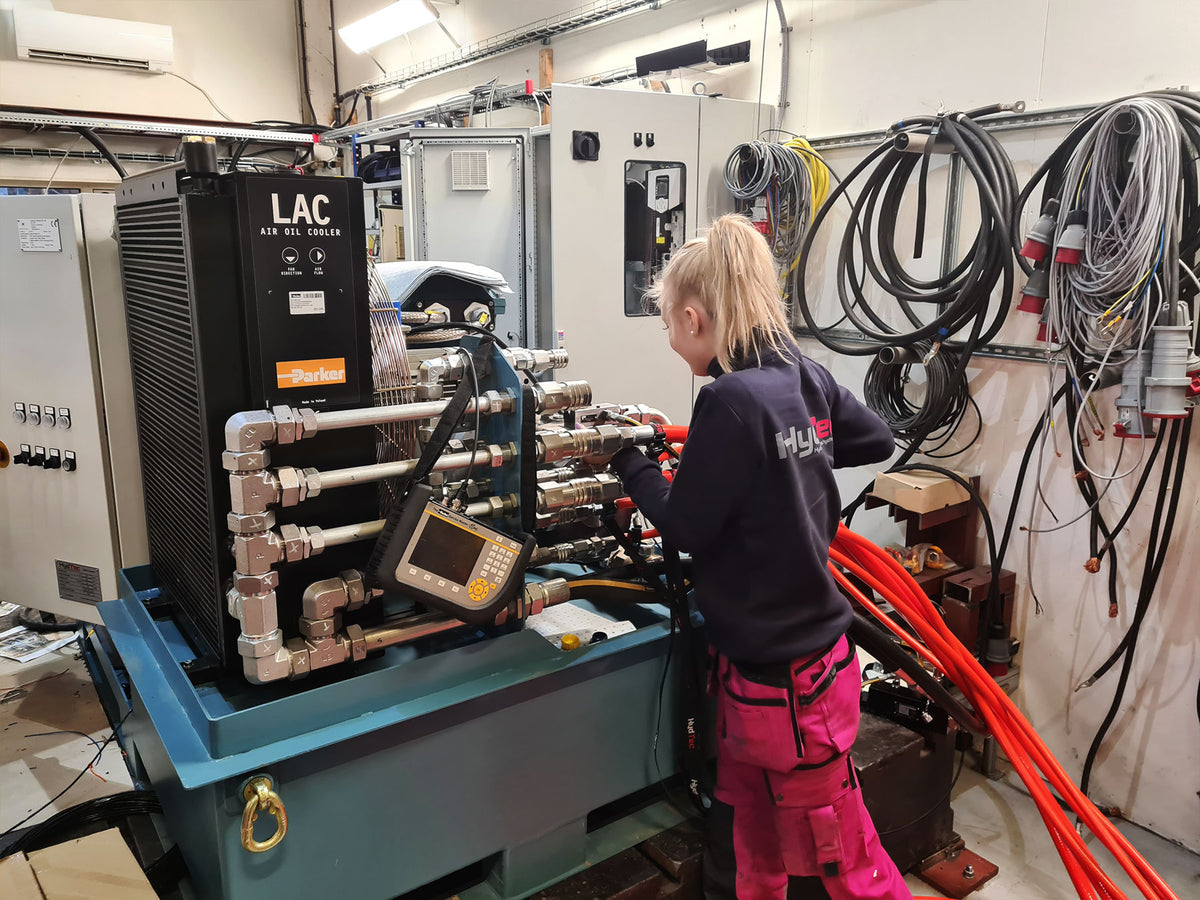 A woman in a workshop stands beside a large hydraulic machine, with coiled hoses and industrial equipment visible in the background.