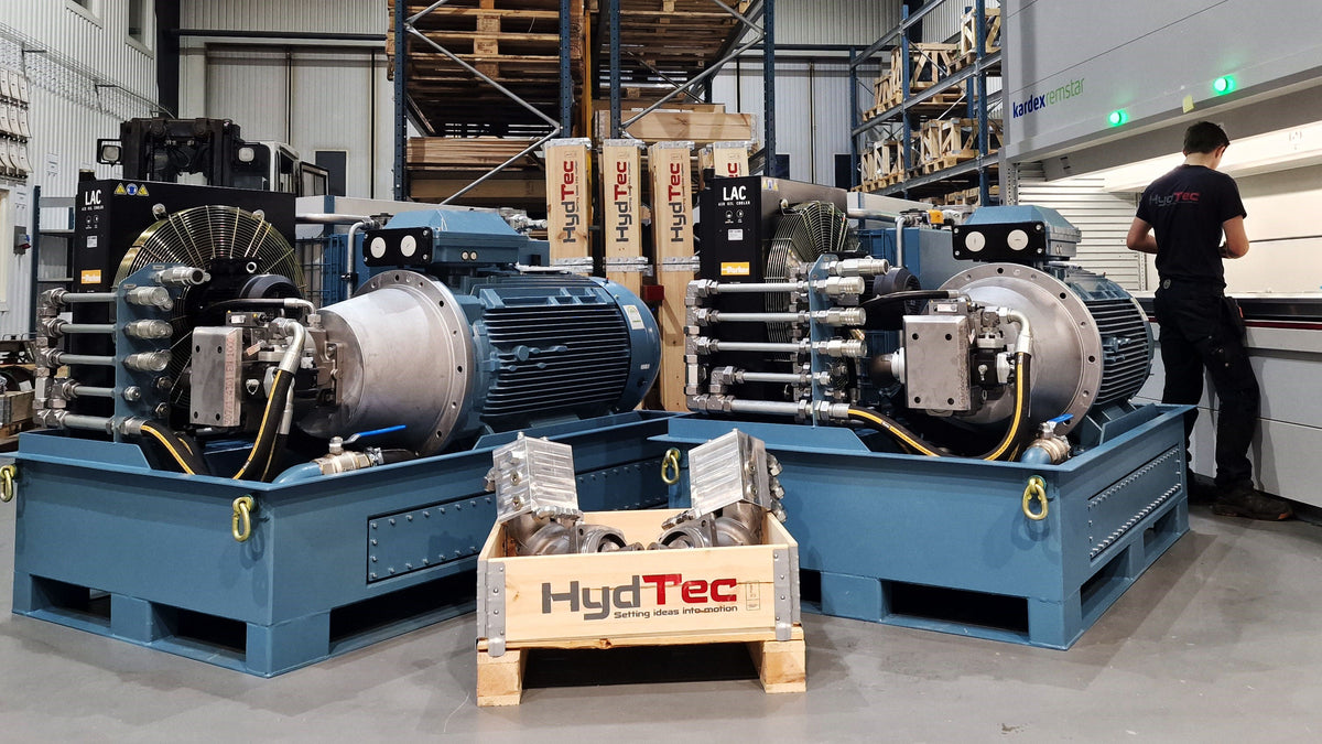 Two large HydTec hydraulic power units on blue metal bases in a warehouse setting, with a branded crate in the foreground and a worker standing near the right side.