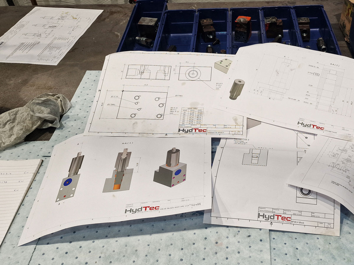 HydTec technical drawings and blueprints spread out on a workbench, alongside a blue parts storage tray containing small mechanical components.